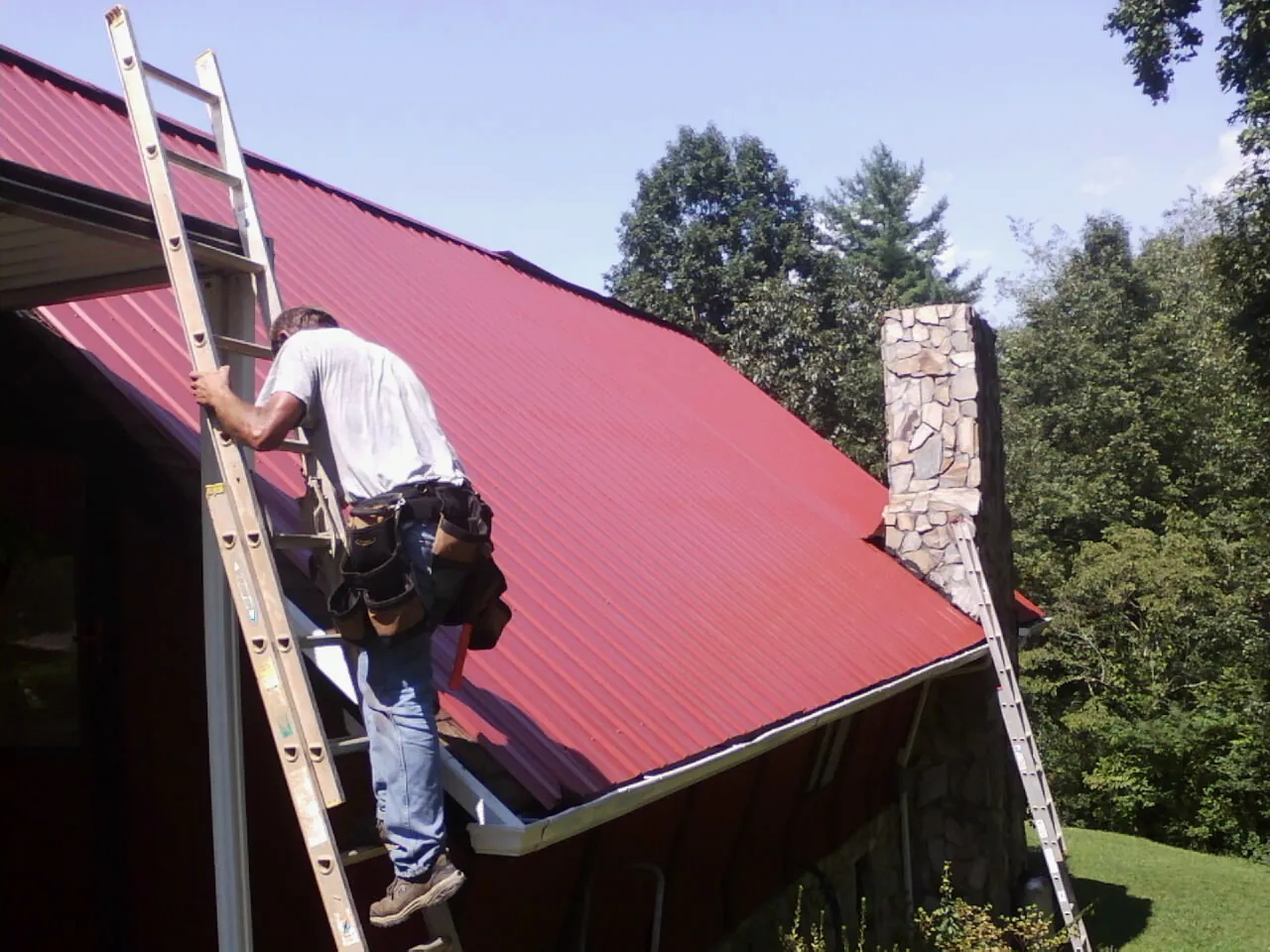 Roofer on a ladder beside a red metal roof and stone chimney — Soffit & Fascia Repair by Roofing Brothers in Wisner